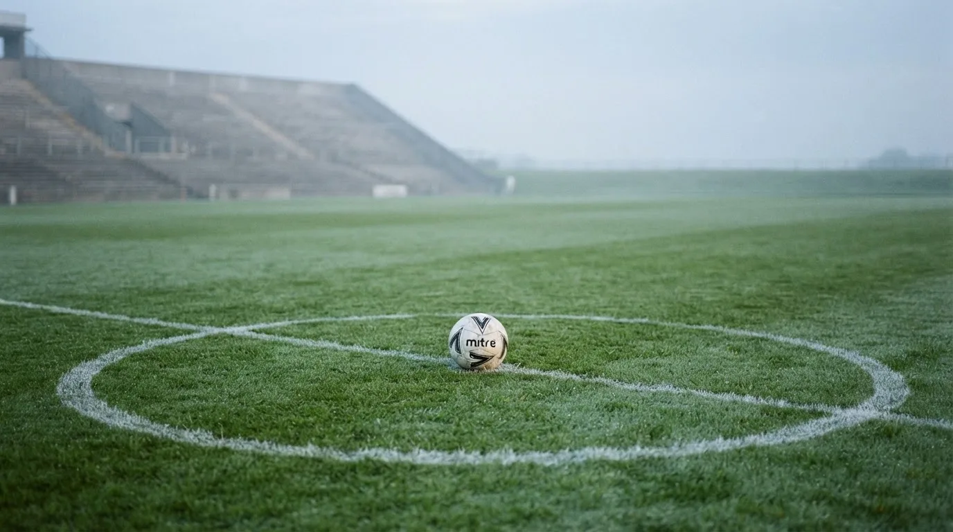 Ein Fußball liegt auf dem Mittelkreis eines leeren Rasenplatzes am frühen Morgen, im Hintergrund eine ferne Tribüne im Dunst.