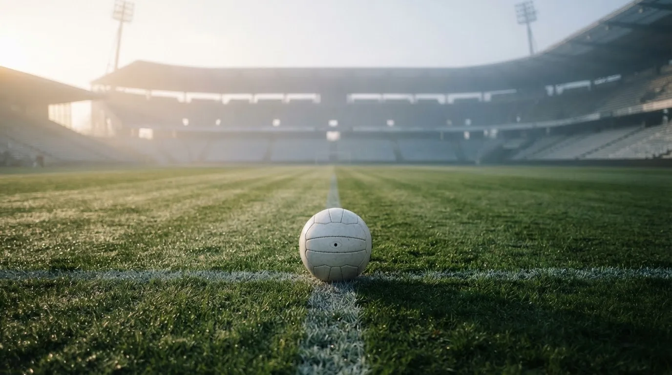 Ein Fußball liegt auf dem Mittelkreis eines leeren Rasenplatzes am frühen Morgen, im Hintergrund eine ferne Tribüne im Dunst.