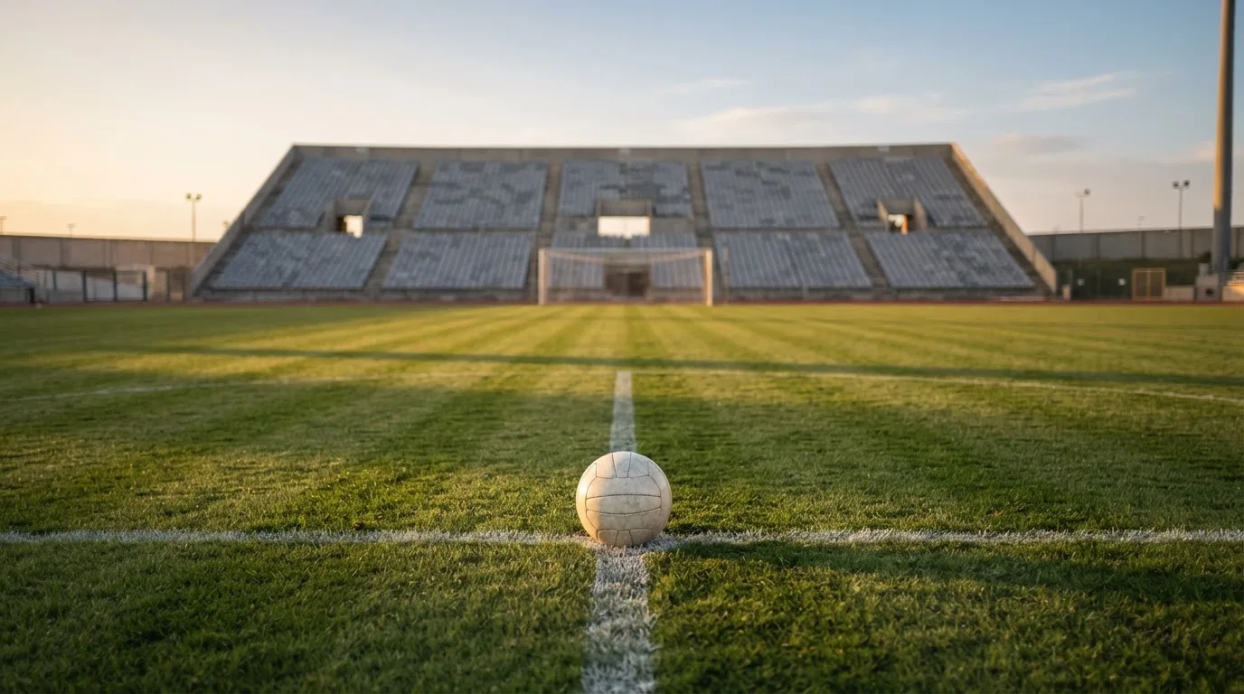 Ein Fußball liegt auf dem Anstoßpunkt eines leeren Rasenplatzes, dahinter eine menschenleere Tribüne im weichen Abendlicht.
