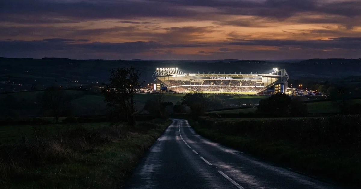 Ein Stadion unter Flutlicht aus großer Entfernung gesehen, davor eine dunkle Landstraße und einzelne Bäume in der Abenddämmerung.
