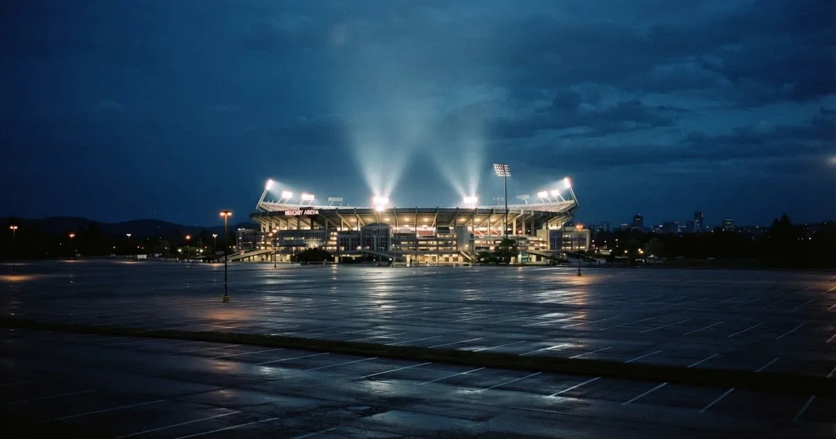 Ein Fußballstadion unter Flutlicht aus großer Entfernung gesehen, die Lichtkegel stehen im dunklen Abendhimmel über leeren Parkplätzen.