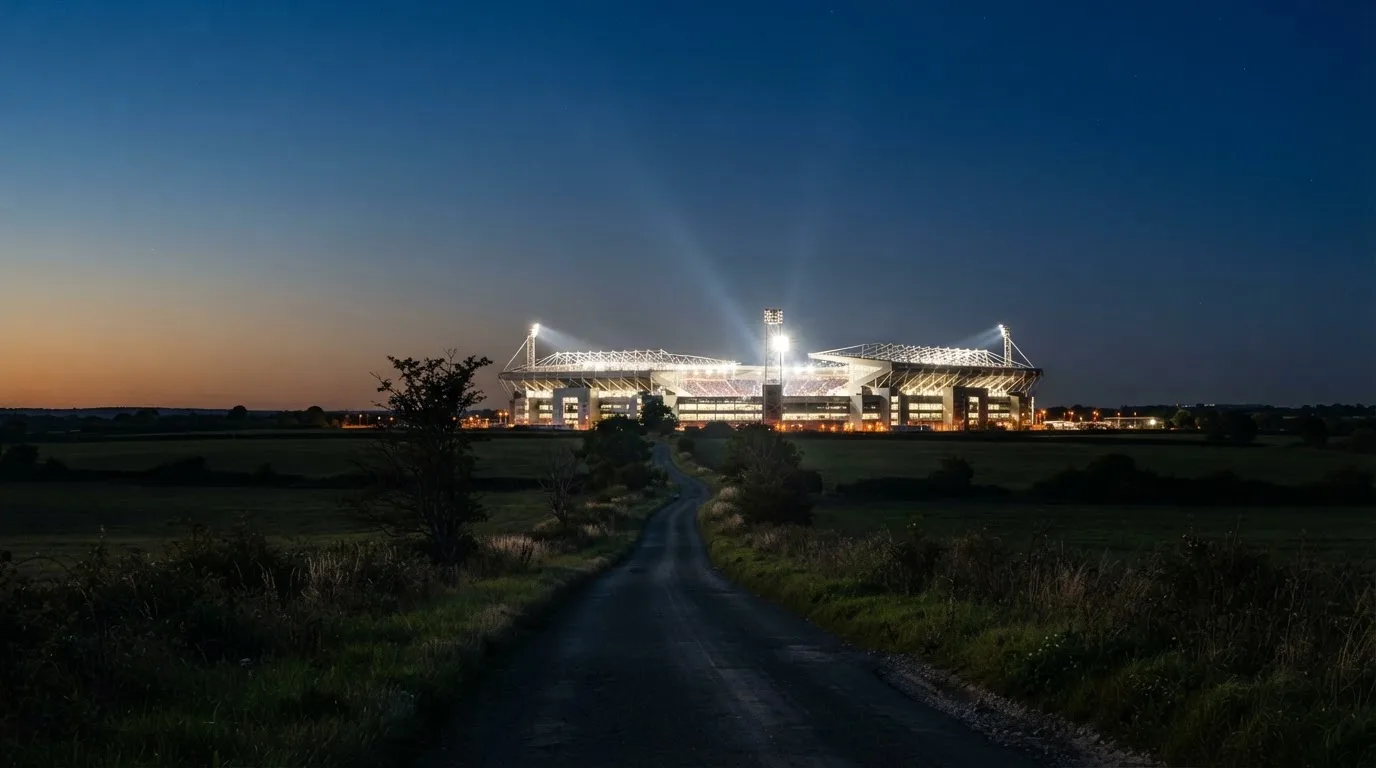 Ein großes Stadion unter Flutlicht aus weiter Entfernung gesehen, davor eine dunkle Landstraße und ein paar Bäume.
