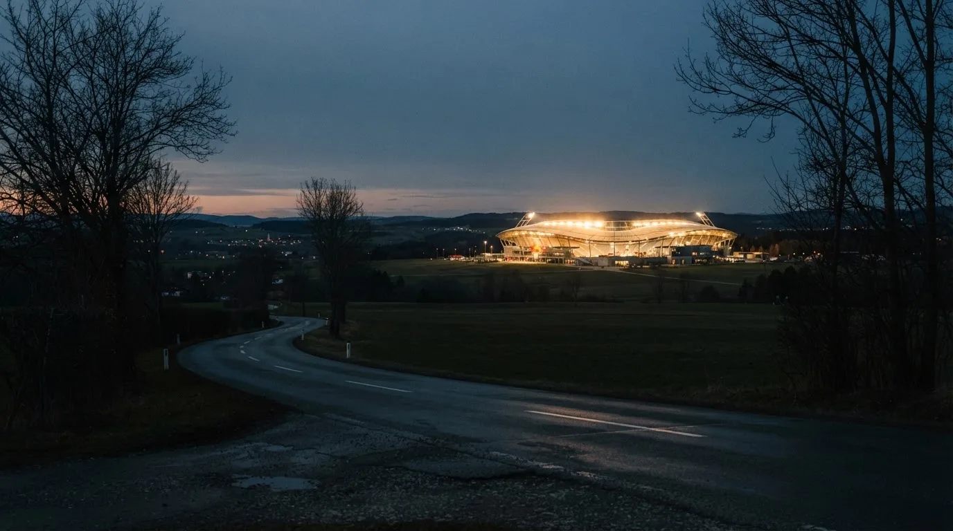 Ein Stadion unter Flutlicht aus großer Entfernung gesehen, davor eine dunkle Landstraße und einzelne Bäume in der Dämmerung.