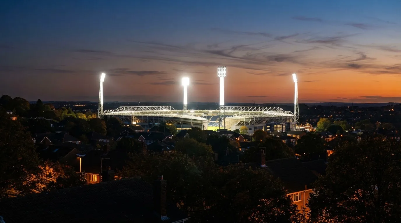 Ein Flutlichtstadion bei Dämmerung aus weiter Ferne gesehen, die Lichtmasten leuchten über Dächern und Bäumen.